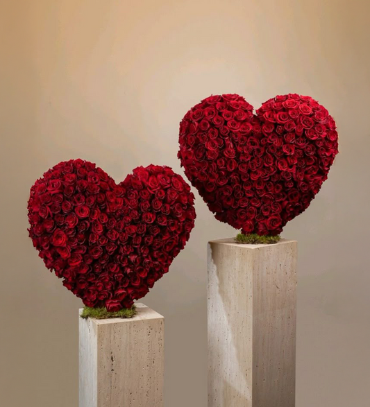 Two heart-shaped arrangements of red roses on stone plinths against a beige background