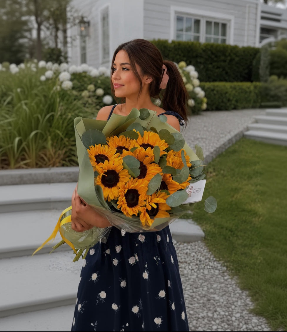 A woman outdoors holding a large bouquet of sunflowers wrapped in green paper. She is wearing a navy dress with a white floral pattern, and a white house with green hedges is in the background.