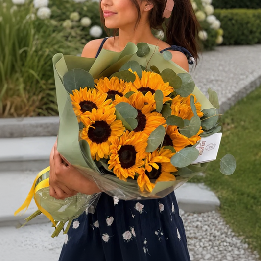 A woman outdoors holding a large bouquet of yellow sunflowers wrapped in green paper. She is wearing a navy blue dress with a white floral pattern, standing on a gravel path with a house and garden in the background.