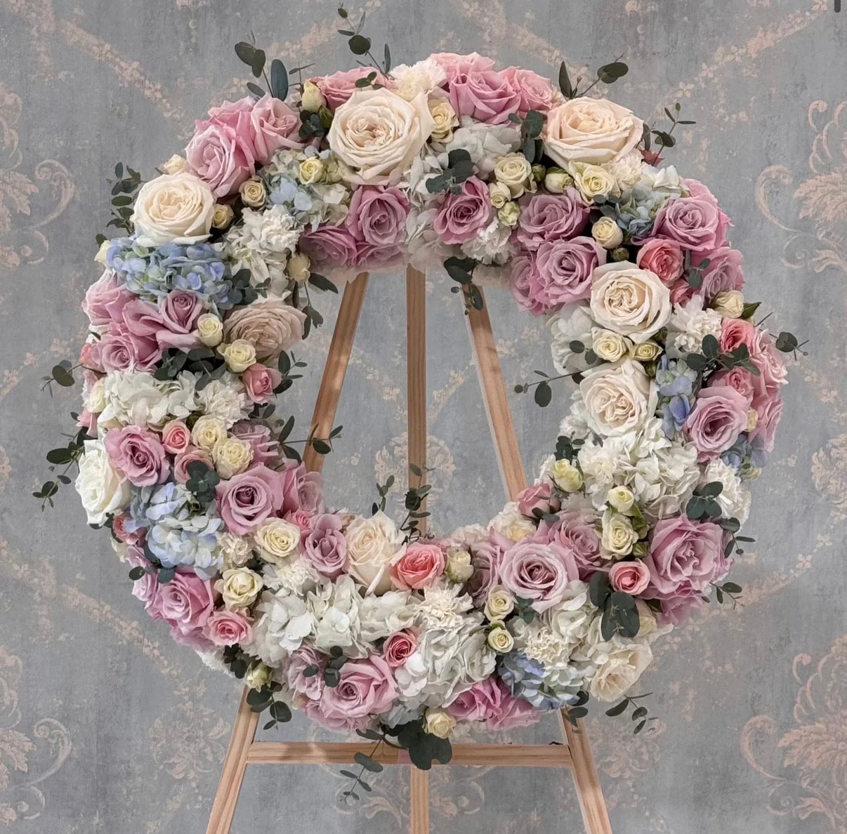 Floral wreath with pink, white, and purple flowers on a stand against a decorative wall.