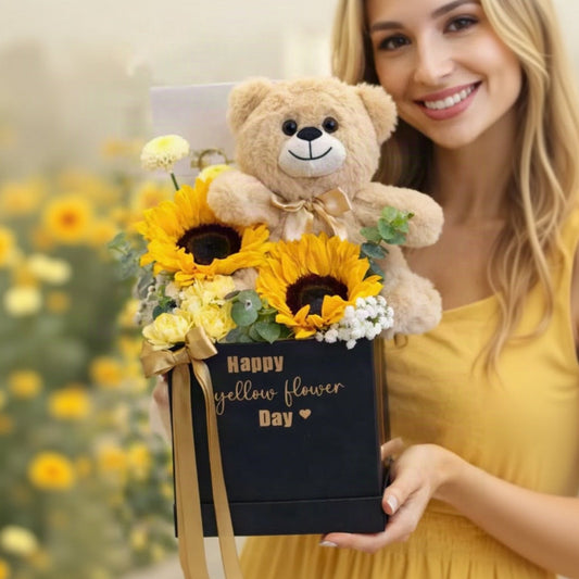 A smiling woman in a yellow dress holds a black gift box decorated with a gold ribbon and the words 'Happy yellow flower Day'. The box is filled with bright yellow sunflowers, yellow and white flowers, and topped with a large beige teddy bear wearing a small gold bow.