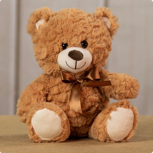A brown plush teddy bear with a white snout and paw pads, sitting on a beige surface against a soft gray background. The bear wears a brown satin bow around its neck.