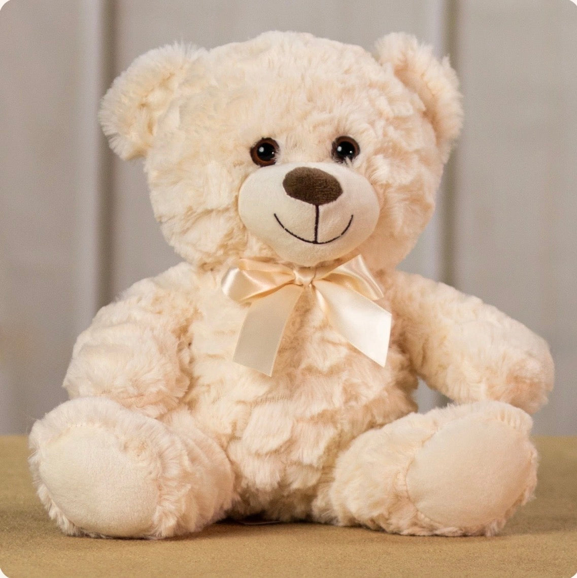 A cream-colored plush teddy bear with a cream satin bow around its neck, sitting on a beige surface against a soft neutral background.