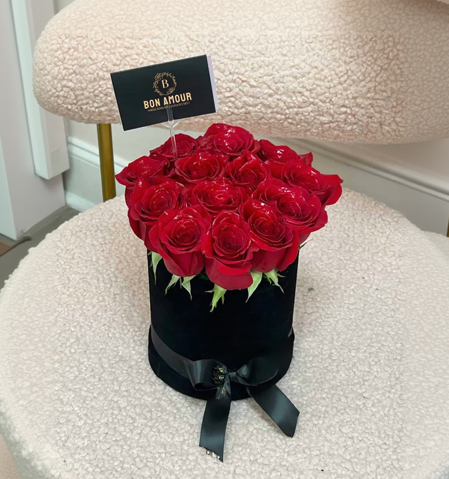 A round black gift box filled with red roses, placed on a cream-colored textured chair. The box is tied with a black ribbon and has a small card that reads 'BON AMOUR'.