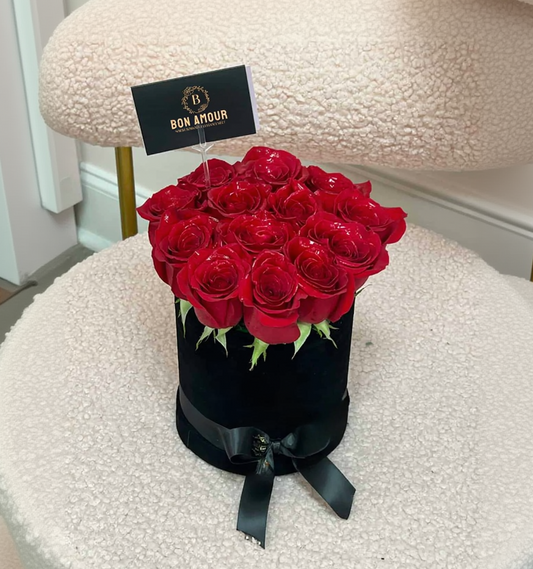 A round black gift box filled with red roses, placed on a cream-colored textured chair. The box is tied with a black ribbon and has a small card that reads 'BON AMOUR'.
